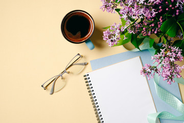 Flat lay home office desk. Female workspace with white paper notebook, glasses, coffee cup and lilac flowers on beige background. Top view feminine background. Fashion blogger desktop concept.