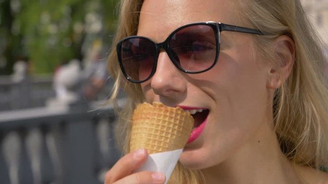 SLOW MOTION, HANDHELD, CLOSE UP, DOF: Girl with sunglasses eating delicious ice cream on a hot summer day. Cheerful blonde woman looking around the city while eating a cone of chocolate ice cream.