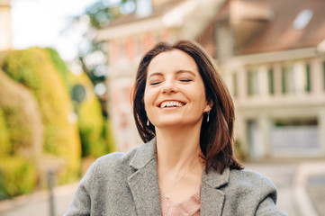 Outdoor portrait of beautiful woman wearing grey coat, laughing, eyes closed