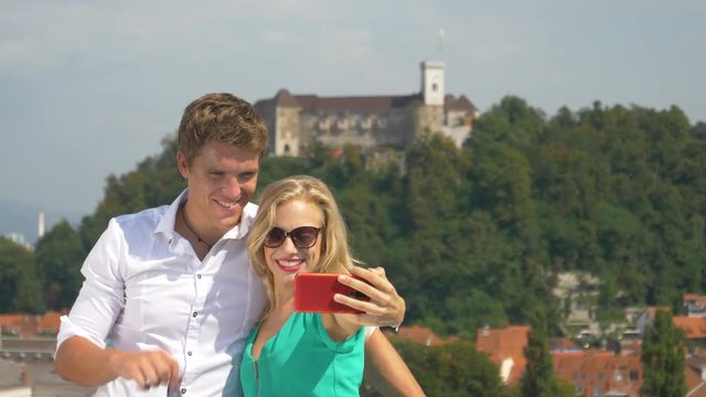 CLOSE UP, DOF: Excited Caucasian guy gives the thumbs up during a travel vlog with his beautiful girlfriend. Cheerful couple taking pictures together from a rooftop in sunny Ljubljana city center.