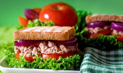 Tuna sandwiches on a square china dish, tomatoes, lettuce, red onion. Selective focus. Two fish sandwiches. White-green background, cloth napkins.