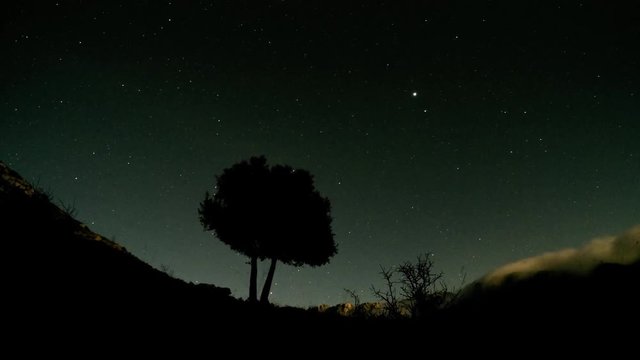 Time lapse of stars with the Big Dipper, the polar star, a mediterranean tree, and  the Sierra Nevada mountain range covered in clouds with the Cerro del Espinar and  Los Castillejos, in Spain.