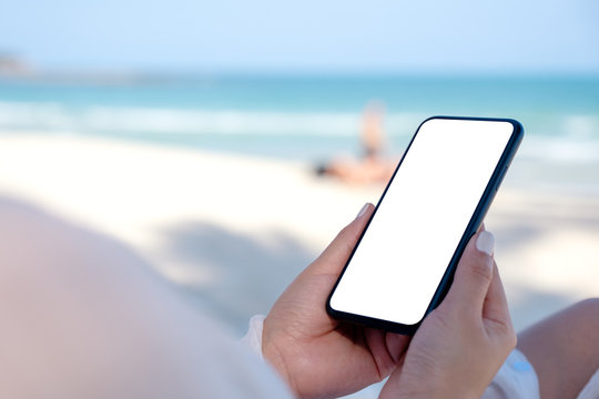 A woman holding white mobile phone with blank desktop screen with sea and blue sky background on the beach