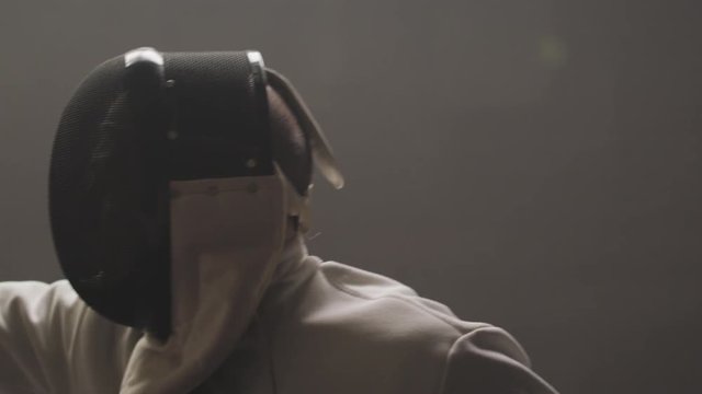Close-up silhouette of a fencers epee mask in a dark and dusty gym in slow motion