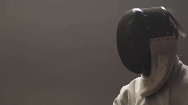 Close-up silhouette of a fencers epee mask in a dark and dusty gym in slow motion