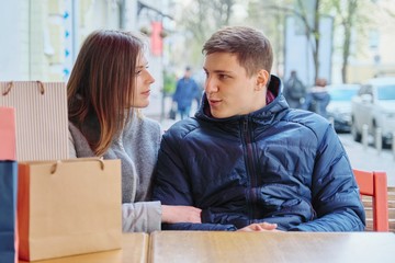 Young talking couple with shopping bags in street cafe, waiting for cup of coffee and tea