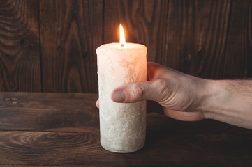 A hand is holding a big white burning candle. On wooden background