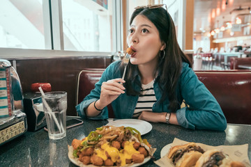 young cheerful asian woman in restaurant diner eating hamburger and chicken wings by fork enjoy...