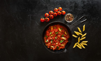 Penne pasta with tomato sauce and green basil decorated in a iron pan on a black background. Top view with copy space
