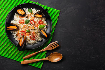 Fried rice with seafood mussels, shrimps, basil in a black plate with wooden spoon and fork on green bamboo mat and stone table