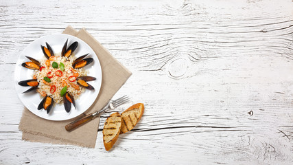 Fried rice with seafood mussels, shrimps and basil in a plate with burlap, toasted baguette and fork on white cracked wooden table