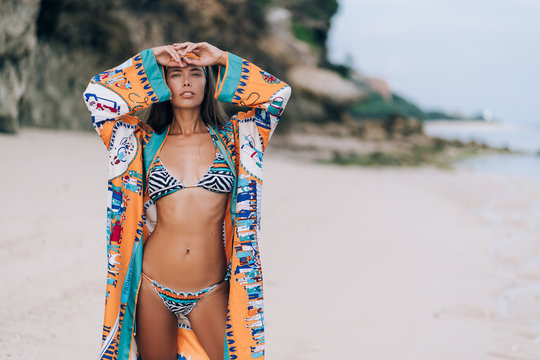 Beautiful sexy girl in swimwear walking on sandy beach with rock on background