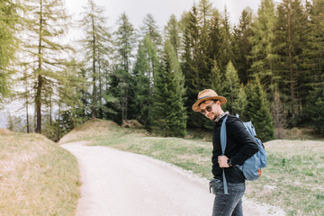 Outdoor portrait of handsome stylish guy thoughtfully standing on the road, resting after long hiking. Male traveler wearing trendy hat and sunglasses posing on forest background enjoying trip.