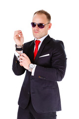 Portrait of elegant and stylish business man in black suit, red tie, white shirt and black sunglasses, adjusts his cufflinks - isolated on white background. Leather bracelet and clock on man's wrist