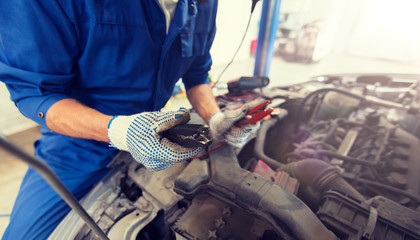 car service, repair, maintenance and people concept - auto mechanic man with cleats charging battery at workshop