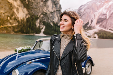 Naklejka premium Portrait of excited woman touching her hair and looking away with smile walking near lake. Pretty young lady in black jacket arrive to look at mountains on vacation and standing in front of blue car.