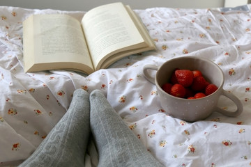 Feet in knit grey socks, bowl of strawberries and book on a bed. Unrecognizable person enjoying leisure time. Selective focus.