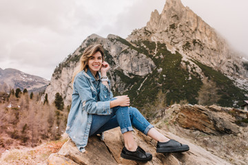 Naklejka na ściany i meble Inspired smiling girl wearing black leather shoes chilling on stone after long trekking and posing with pleasure. Portrait of adorable female traveler addicted to active tourism in mountains.