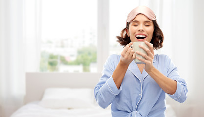 people and bedtime concept - happy young woman in pajama and eye sleeping mask drinking coffee from...