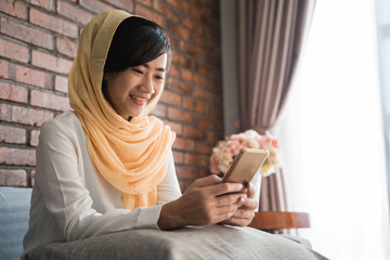 asian muslim woman using mobile phone at home sitting on a couch