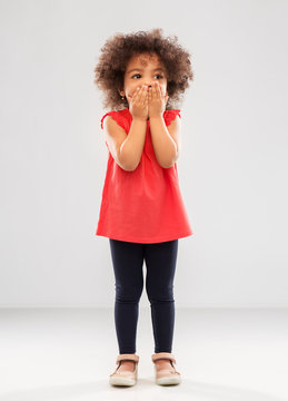 childhood, expression and emotion concept - confused little african american girl covering mouth by hands over grey background