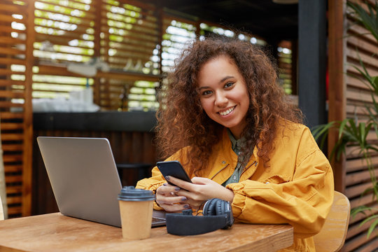Portrait of smiling young beautiful dark skinned curly student girl on a cafe terrace, holding smartphone on his hands, wearing in yellow coat, enjoys the day.