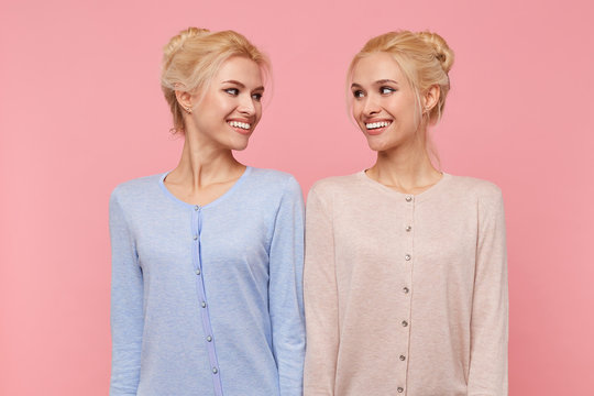 Photo of young broadly smiling happy blonde twins, looking at each other isolated over pink background.