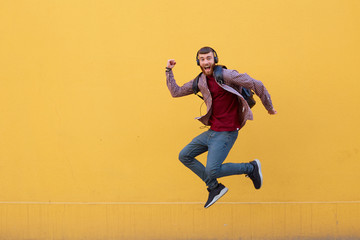 Fototapeta na wymiar Happy young handsome ginger bearded man jumping like superhero, full of energy, wearing in basic clothes with backpack. Looking at the camera with wide open mouth over a yellow wall with copy space.