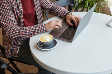 Clouse up in a cafe on a white table, men's hands are working on the keyboard of the laptop, next to a gray cup of coffee.