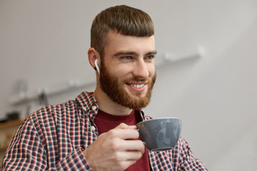 Portrait of a young handsome red-bearded guy broadly smiling and enjoying delicious brewed coffee, wearing in basic clothes.