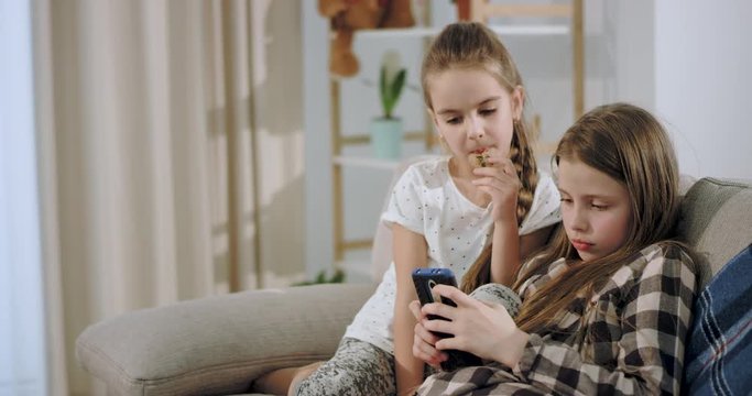 Portrait of a cute two sisters playing on the smartphone concentrated while sitting on the sofa in a spacious living room