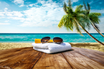 Fototapeta na wymiar Wooden table background of free space for your decoration. White towel background with sunglasses and beach landcsape with palms and ocean. Blue sky with sun light. Summer time on beach. 