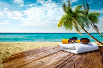 Wooden table background of free space for your decoration. White towel background with sunglasses and beach landcsape with palms and ocean. Blue sky with sun light. Summer time on beach. 