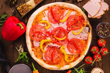 Venice pizza with ham, tomatoes and paprika on a dark wooden background with ingredients around (close top view)