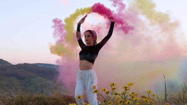 Bellissima ragazza felice, danza in una campagna con prato verde, con dei fumogeni colorati. Video al rallentatore.