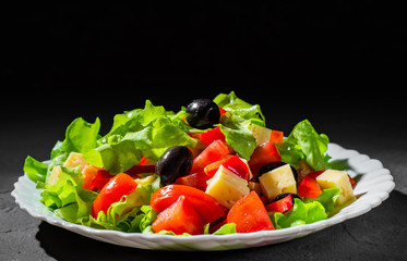fresh vegetables salad with lettuce, olive, pepper, tomato and cheese in white plate on dark black background