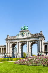 The arcade du Cinquantenaire, the triumphal arch erected by king Leopold II in the Cinquantenaire park in Brussels, Belgium, with a flowerbed in full bloom in the foreground against blue sky.