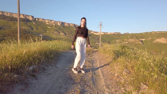 Giovane ragazza con maglia nera e tuta grigia balla con musica allegramente in una strada sterrata, in cima a una collina in campagna. Tra la natura. Tramonto sullo sfondo.