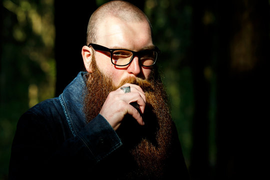 Vape bearded man. An adult white short-haired man with a very long beard in glasses smokes an electronic cigarette outside in the dark forest. Bad habit that is harmful to health. - Powered by Adobe
