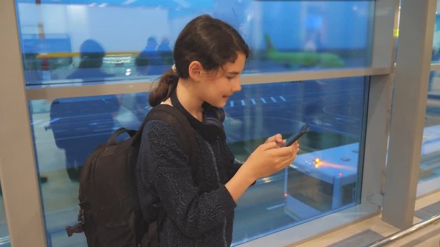 daughter with a backpack airplane flights airport concept. girl teenager with a smartphone looks out the window at the airport area airplanes and bus at a distance. teen girl looks at the schedule