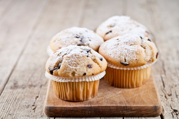 Four homemade fresh muffins with sugar powder and blueberries on cutting board on rustic wooden table.