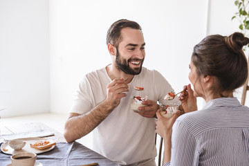 Image of joyous brunette couple eating panna cotta dessert together while sitting at table at home