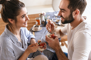 Image of caucasian brunette couple eating panna cotta dessert together while sitting at table at home