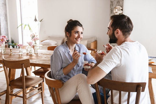 Image of cheerful brunette couple eating panna cotta dessert together while sitting at table at home