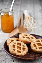 Fresh baked tarts with marmalade or apricot jam filling and on ceramic plate and kitchen bakery utensil on on rustic wooden table.