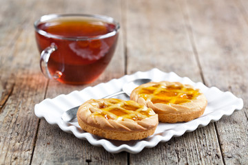 Fresh baked tarts with marmalade or apricot jam filling on white ceramic plate and cup of tea on rustic wooden table.