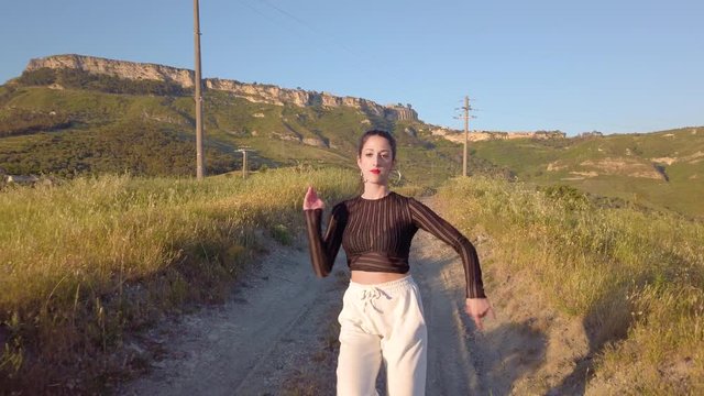 Giovane ragazza con maglia nera e tuta grigia balla con musica allegramente in una strada sterrata, in cima a una collina in campagna. Tra la natura. Tramonto sullo sfondo.