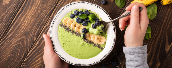 Breakfast detox green smoothie bowl from banana and spinach on wooden background. Healthy food concept with women hands and copy space, top view, banner