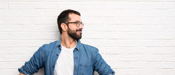 Handsome man with beard over white brick wall posing with arms at hip and smiling