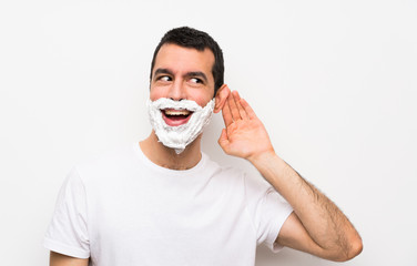 Man shaving his beard over isolated white background listening to something by putting hand on the ear